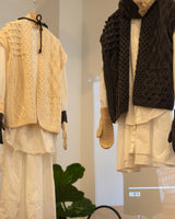 Two styled knit outerwear pieces displayed on hangers against a bright indoor background. The left piece is a cream cable-knit sleeveless vest layered over a white shirt and white pants, with beige gloves hanging at the sides.