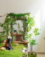 A whimsical mossy forest display featuring knitted stuffed animals, including a red panda and a bird, arranged around a wooden mirror and framed by lush faux greenery and hanging plants.