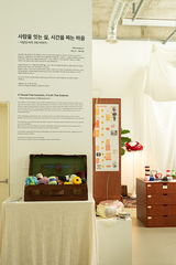 An interior exhibition or gallery scene with printed text panels, a brown open trunk filled with colorful thread-like items, a wooden cabinet, and draped white fabric in a bright indoor space.