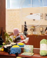 A cozy indoor scene showing a wooden shelf with colorful textured pot covers, a ball of light green Dandi yarn, and a postcard. The background is softly blurred with warm lighting and decorative objects, and a large white line-art graphic with Korean text overlays the upper portion.