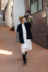 A woman walking down an outdoor alley wearing a dark navy textured cardigan with patterned knit sleeves, a white midi dress, olive green socks, and a tan baseball cap.